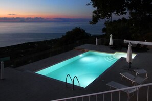 Ein rechteckiger Pool mit hellem Blauton, umgeben von einer dunklen Liegefläche. Zwei weiße Sonnenschirme und mehrere Liegestühle sind vorhanden.