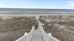 Eine Holztreppe führt zu einem Strand mit einem sandigen Pfad, einer kleinen Holzhütte mit Stühlen und einem klaren Blick auf das Meer.