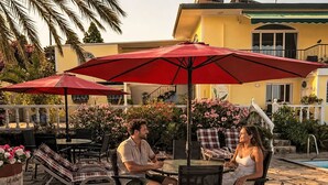 Een terras buiten met een stenen vloer, rode parasols en lounge fauteuils.