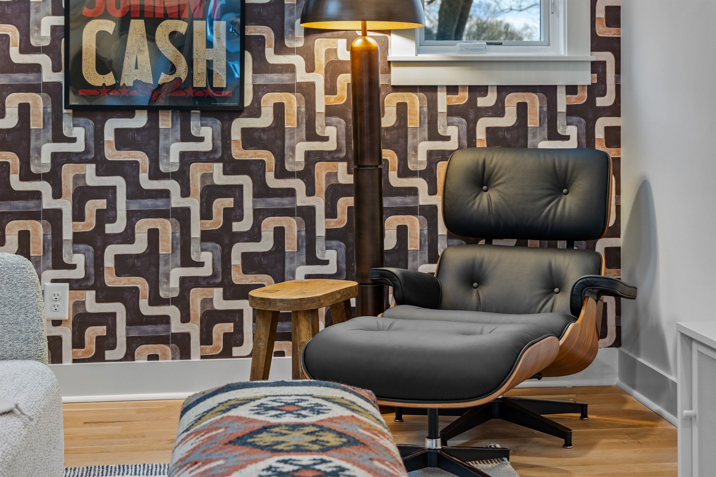 A modern living room with a geometric patterned wallpaper, a black leather chair, a small wooden stool, and a colorful rug.
