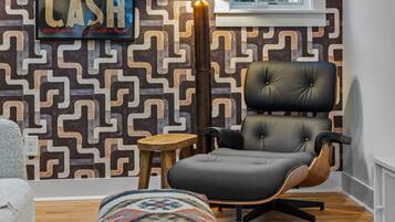 A modern living room with a geometric patterned wallpaper, a black leather chair, a small wooden stool, and a colorful rug.