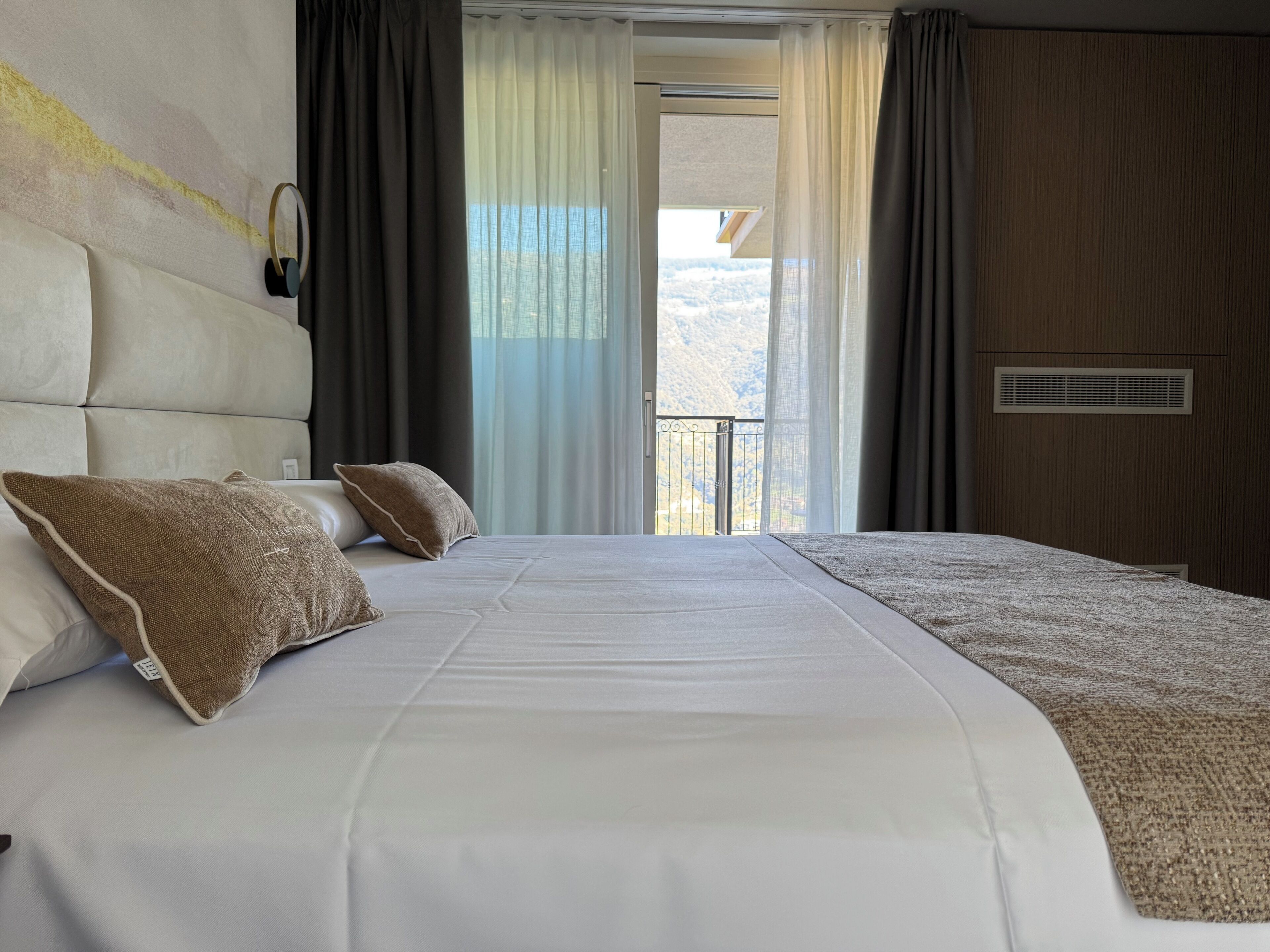 A hotel room with a large bed, two pillows, a headboard, a window with curtains, and a view of a mountainous landscape.