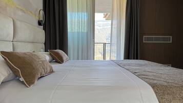 A hotel room with a large bed, two pillows, a headboard, a window with curtains, and a view of a mountainous landscape.