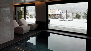 Een modern interieur met een glazen zwembad, zithoek en grote ramen met uitzicht op een besneeid landschap.