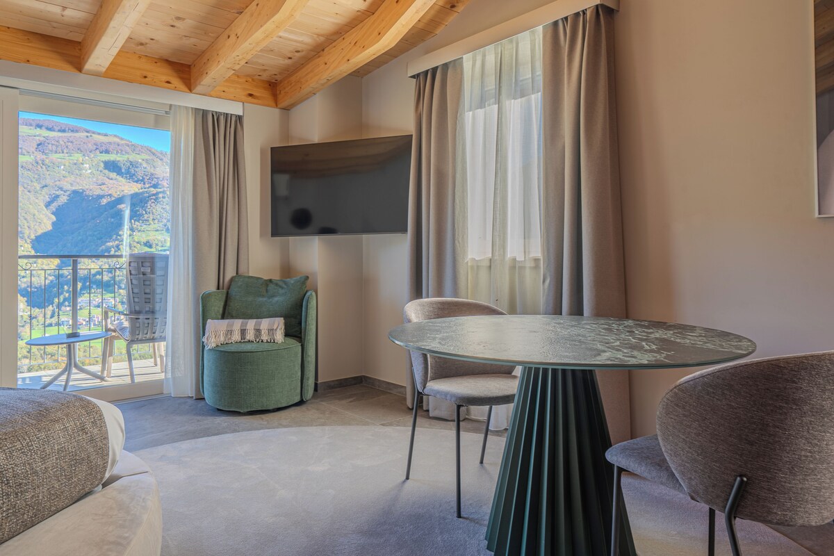 A room with a balcony, a round table, two chairs, a green armchair, and a flat-screen TV mounted on the wall.