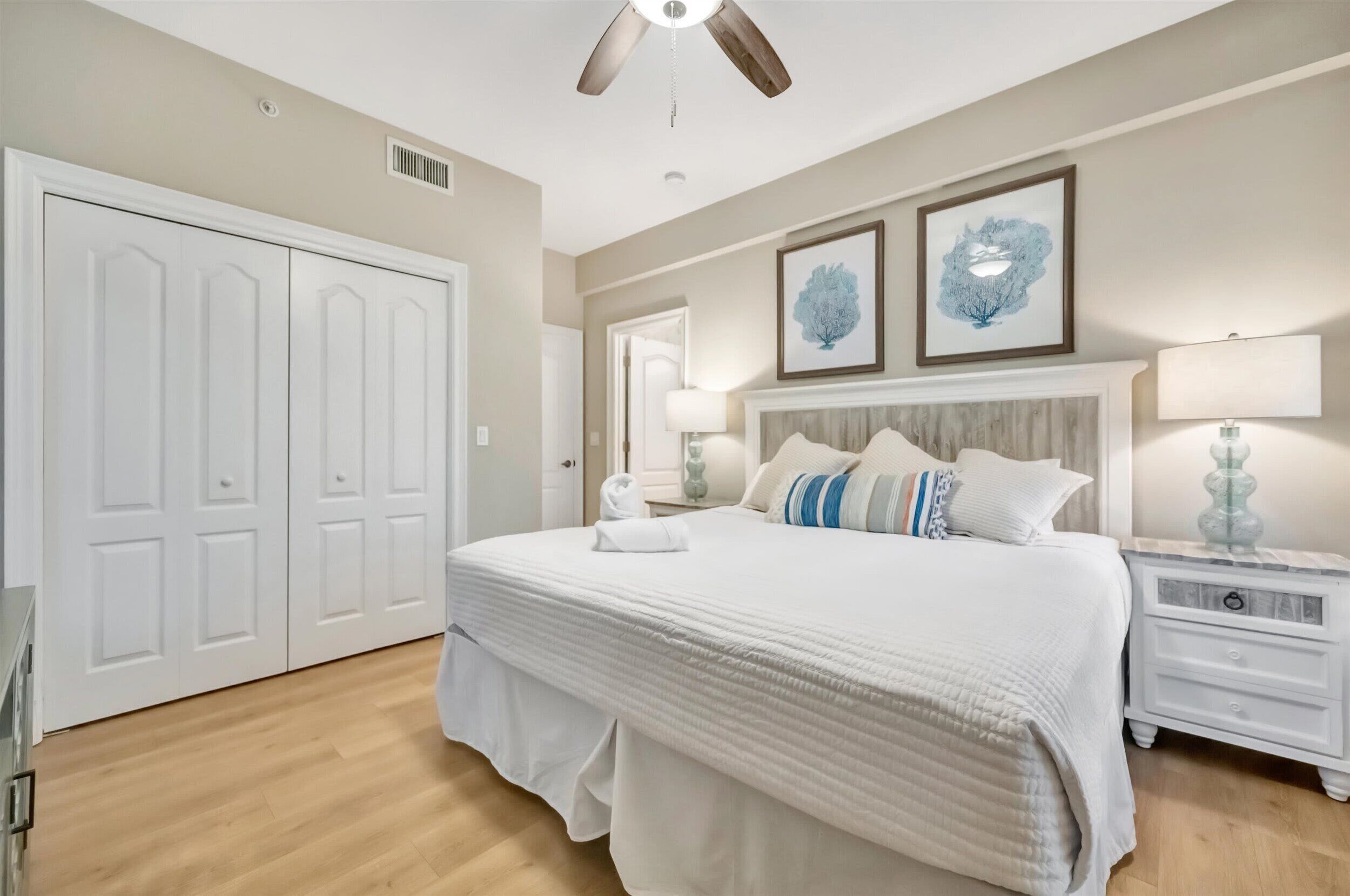 A bedroom with a large bed, a ceiling fan, a nightstand with a lamp, and a door with a mirror.