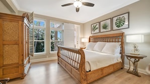Un dormitorio con una cama de madera, un ventilador de techo, una ventana con contraventanas y una obra de arte con plantas enmarcada en la pared.