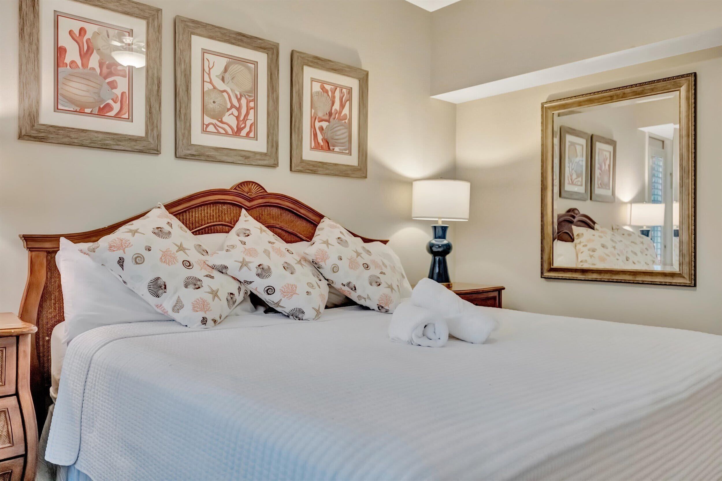 A neatly made bed with a white comforter and pillows with a sea shell pattern, a bedside lamp, and framed artwork on the wall.