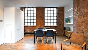 A dining area with a brick wall, large windows, a table with chairs, and a wicker chair. - Spacious Studio in a Converted Warehouse Shoreditch (London)