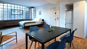 A modern room with a wooden floor, a black table with blue chairs, a bed with white linens, and a large window with a view of buildings. - Spacious Studio in a Converted Warehouse Shoreditch (London)