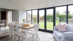 A dining area with a wooden table and white chairs, a sofa with cushions, and a view of a garden through large windows. - Victorian House Sleeps 14 Pool and Gardens (Chichester)