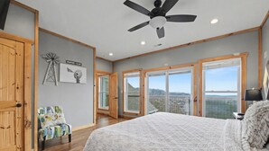 Quarto com cama grande, piso de madeira, ventilador de teto e amplas janelas com vista para o exterior.