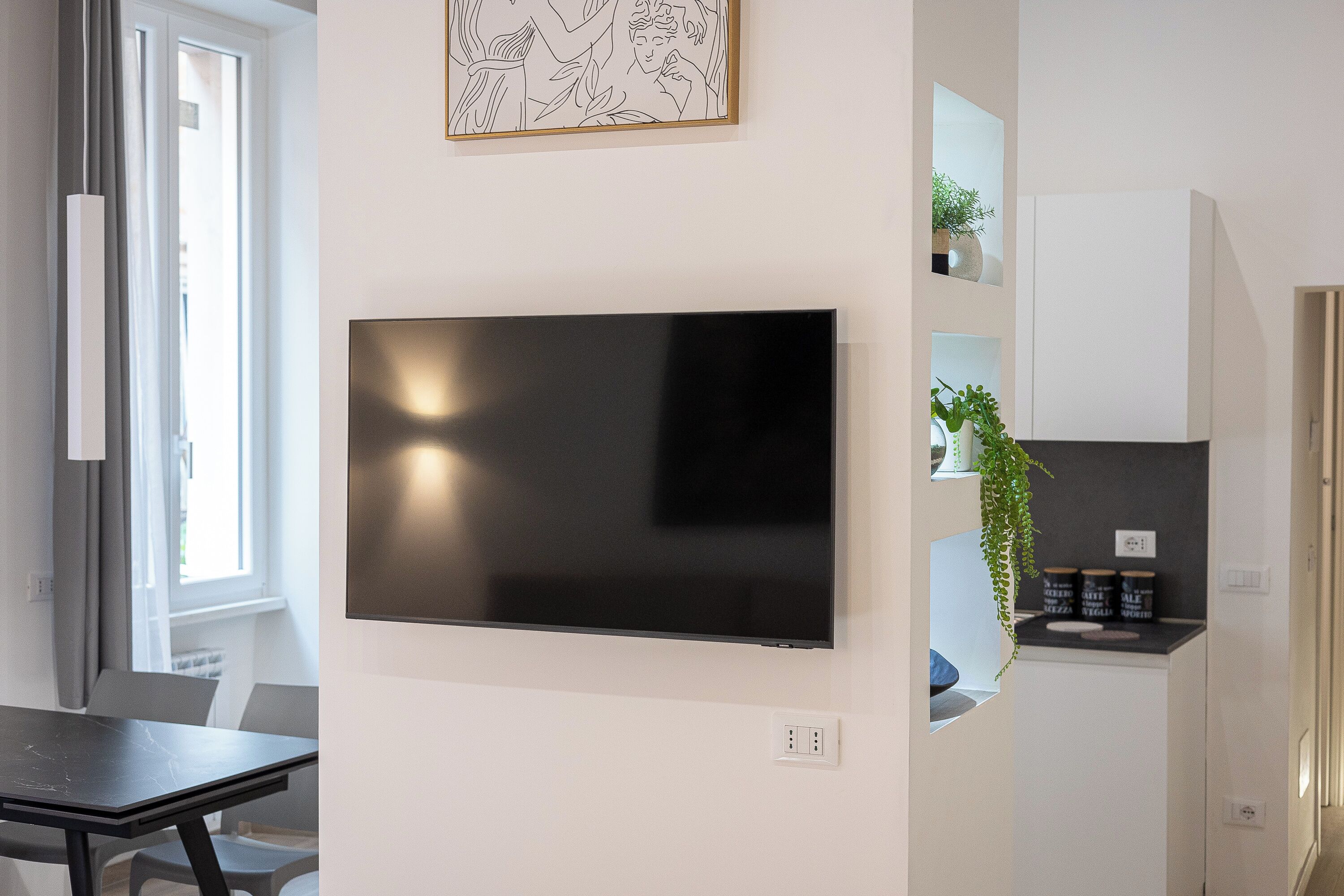 A modern living space with a flat-screen TV mounted on the wall, a dining area with a table and chairs, a kitchen area with a countertop and cabinets, and a window with curtains.