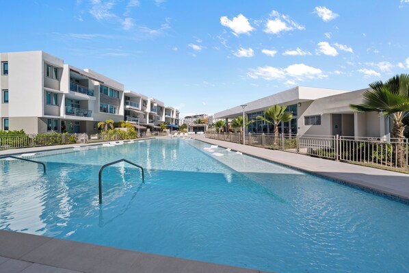 A modern hotel with a swimming pool, multiple balconies, and a clear blue sky.