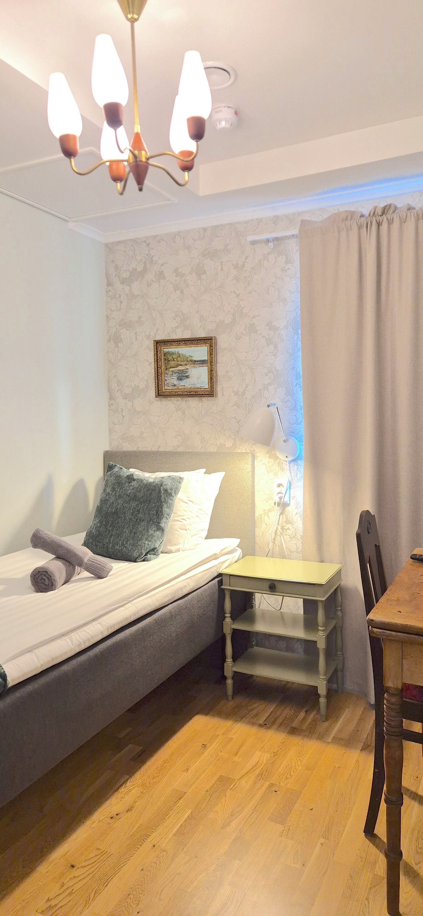 A bedroom with a bed, a bedside table, a chair, a desk, a chandelier, a painting on the wall, and a curtain.