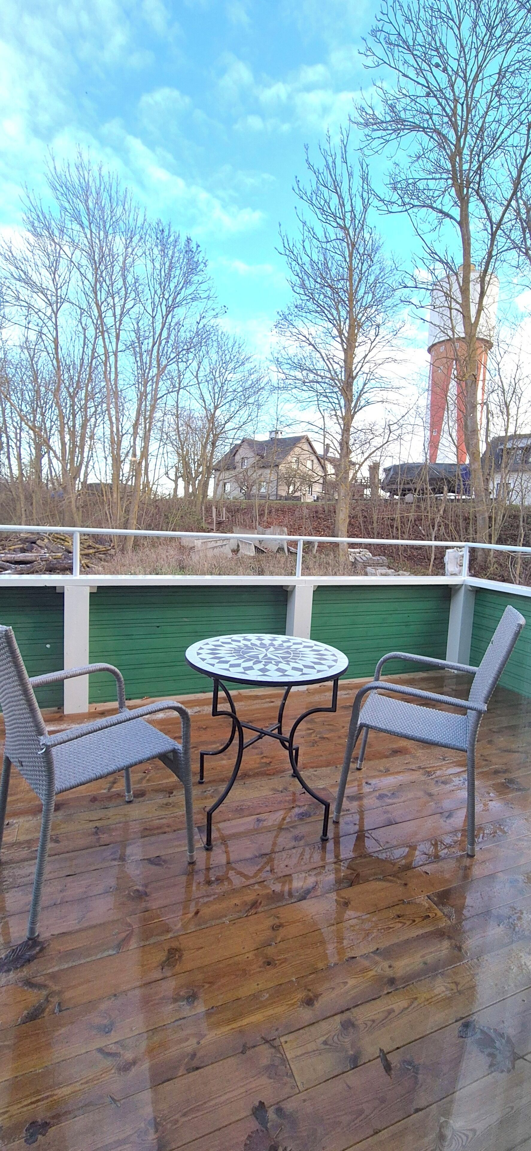 A wooden deck with a round table and two chairs, overlooking a body of water and buildings in the background.