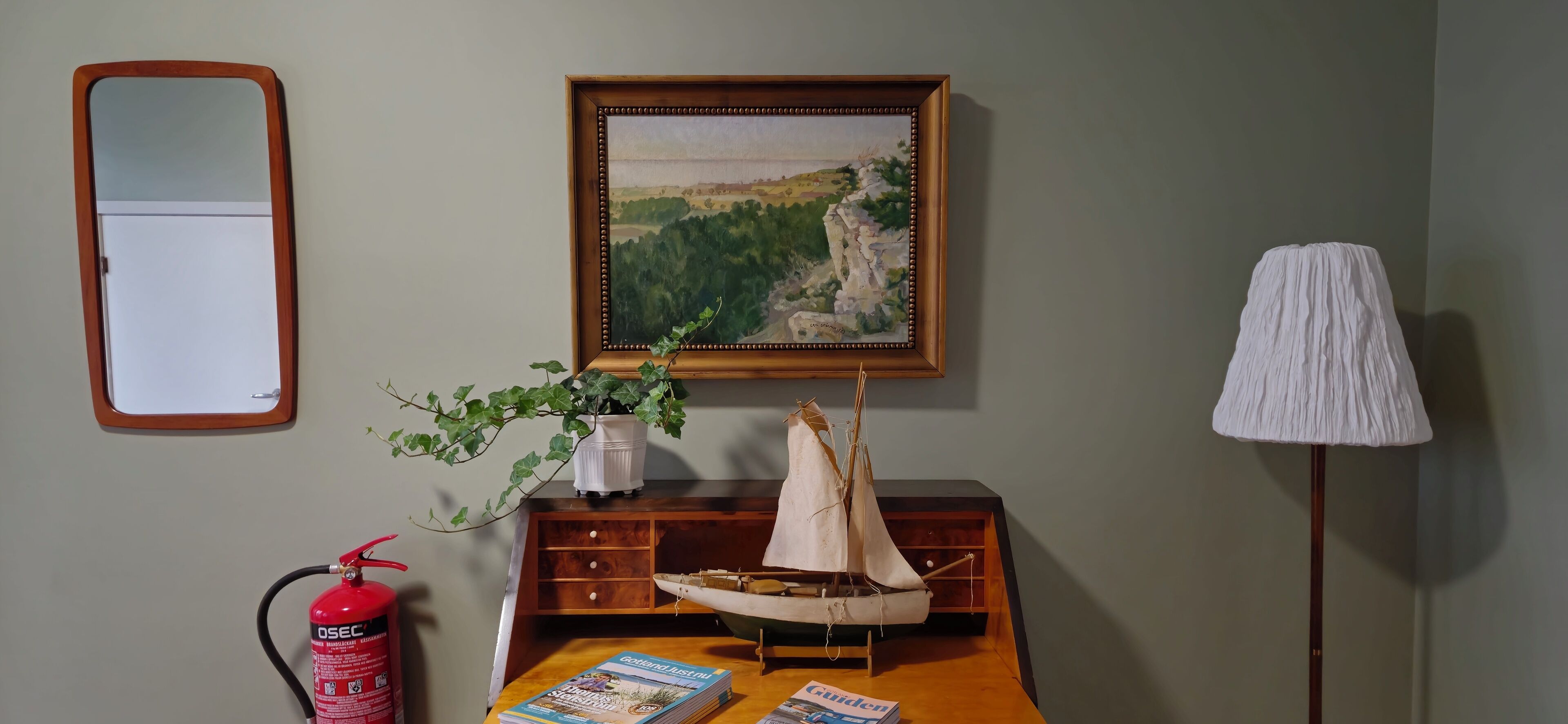Ein Zimmer mit einem Holztisch, einem gerahmten Landschaftsbild, einem Topfpflanzen, einem kleinen Segelbootmodell, einem Feuerlöscher und einer Stehlampe.