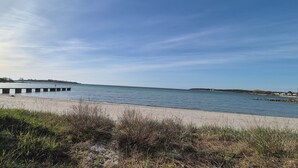 A beach with a pier extending into calm waters, surrounded by grassy areas and distant buildings. - Slitebadens Annex (Slite)