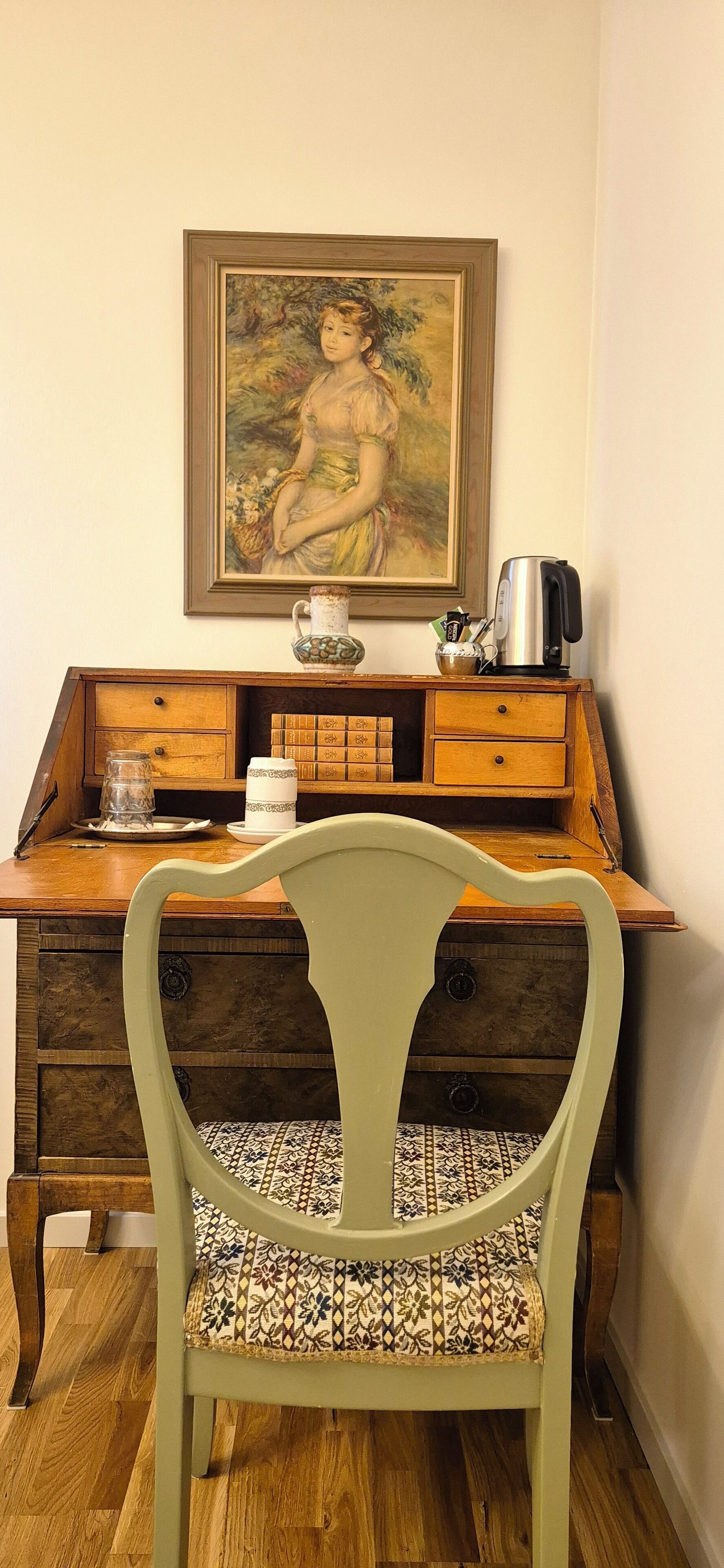 Un escritorio de madera antiguo con cajones, una silla estampada, un cuadro en la pared y una tetera sobre el escritorio.