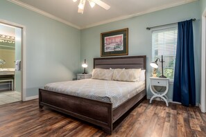 A bedroom with a large bed, a nightstand with a lamp, a framed picture on the wall, and a window with curtains. - Dauphin Luv - Beach Club #201 4 Bedroom Condo (Dauphin Island)