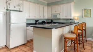 A kitchen with white cabinets, a central island with bar stools, a refrigerator, a microwave, and a coffee maker. - Dauphin Luv - Beach Club #201 4 Bedroom Condo (Dauphin Island)