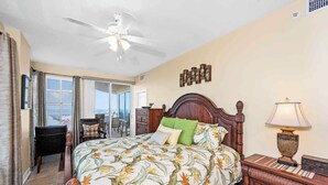 A hotel room with a bed, a nightstand, a ceiling fan, a window with curtains, a chair, and a view of the outside. - Indies 401 | 3BR/3BA Corner Condo | Sleeps 6 | Wraparound Balcony & Views (Gulf Shores)