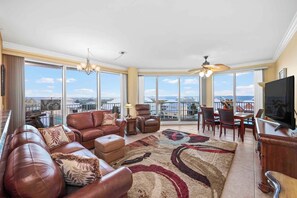 A spacious living room with a large window, leather sofa, and a dining area. - Indies 401 | 3BR/3BA Corner Condo | Sleeps 6 | Wraparound Balcony & Views (Gulf Shores)
