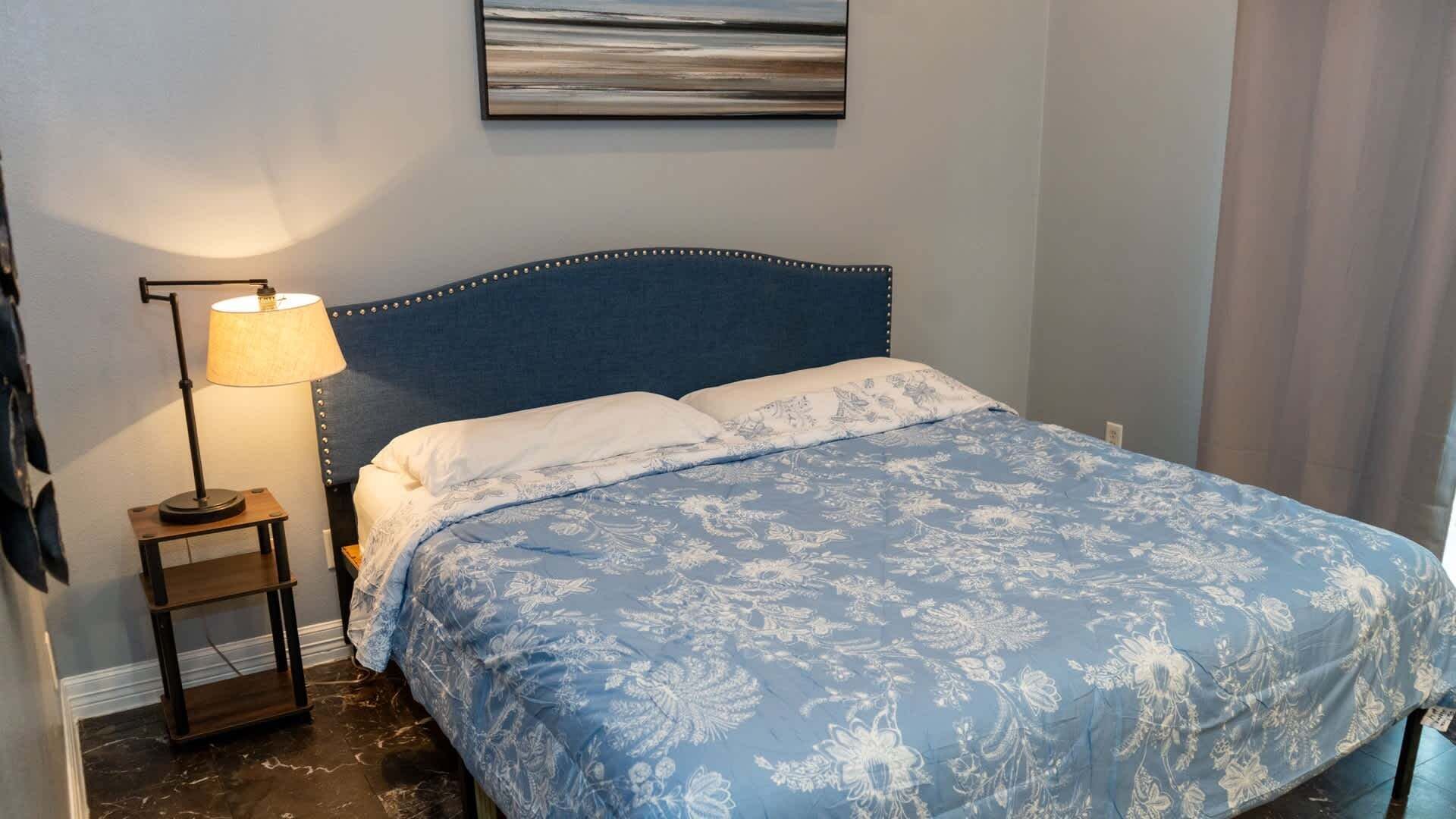 A bedroom with a bed, a nightstand, a lamp, and a painting on the wall.