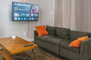 Ein Wohnzimmer mit grauem Sofa, orangefarbenen Kissen, einem Holztisch und einem an der Wand befestigten Flachbildfernseher, der einen Film zeigt.