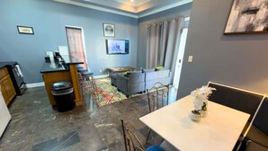 A modern living room with a grey sofa, a dining table, and a kitchen area with a refrigerator and bar stools. - Priced Right Near Beach Well Equipped 2 Bedroom Apts (South Padre Island)