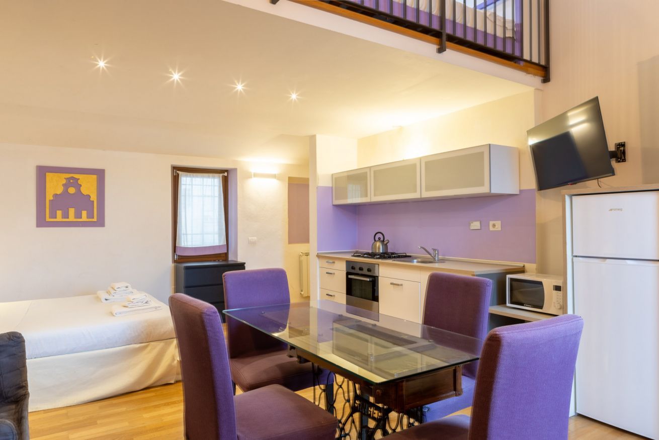 A compact living space with a glass dining table, purple chairs, a kitchenette, a bed, and a TV mounted on the wall.