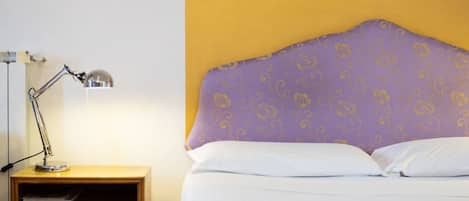 A bed with a purple headboard, a wooden nightstand with a lamp, and a patterned wall.
