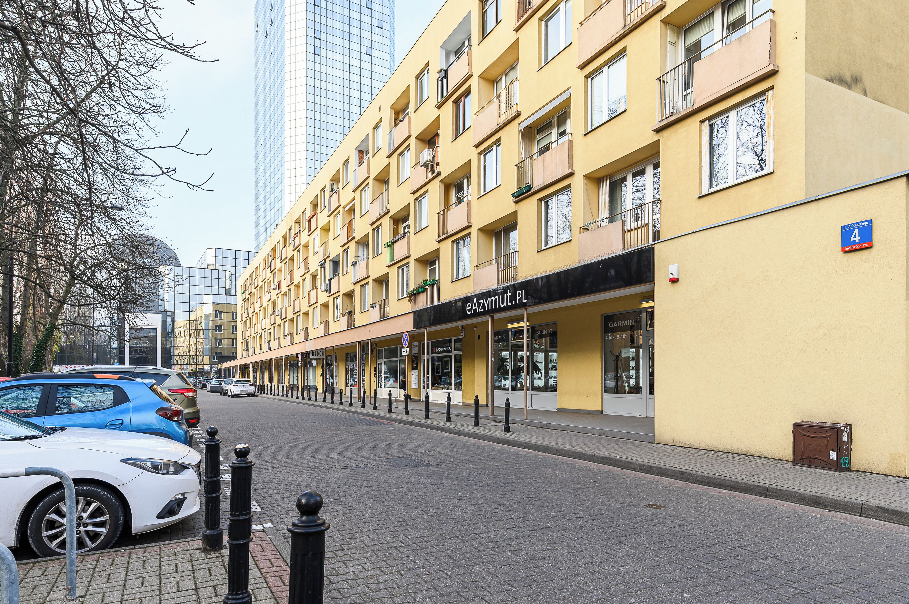 A multi-story yellow building with balconies, a storefront with a sign reading "eAZIMUT.PL", and a parked car on the street.