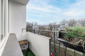 En balkon med udsigt til bygninger og træer, et lille bord og en plantekasse med planter.