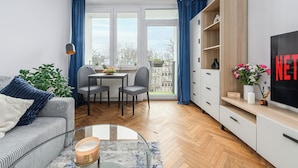 A modern living room with a sofa, coffee table, and a TV. There's a dining area with a table and chairs, and a large window with curtains. - Lumina 2-room apart near the Old Town (Warsaw)
