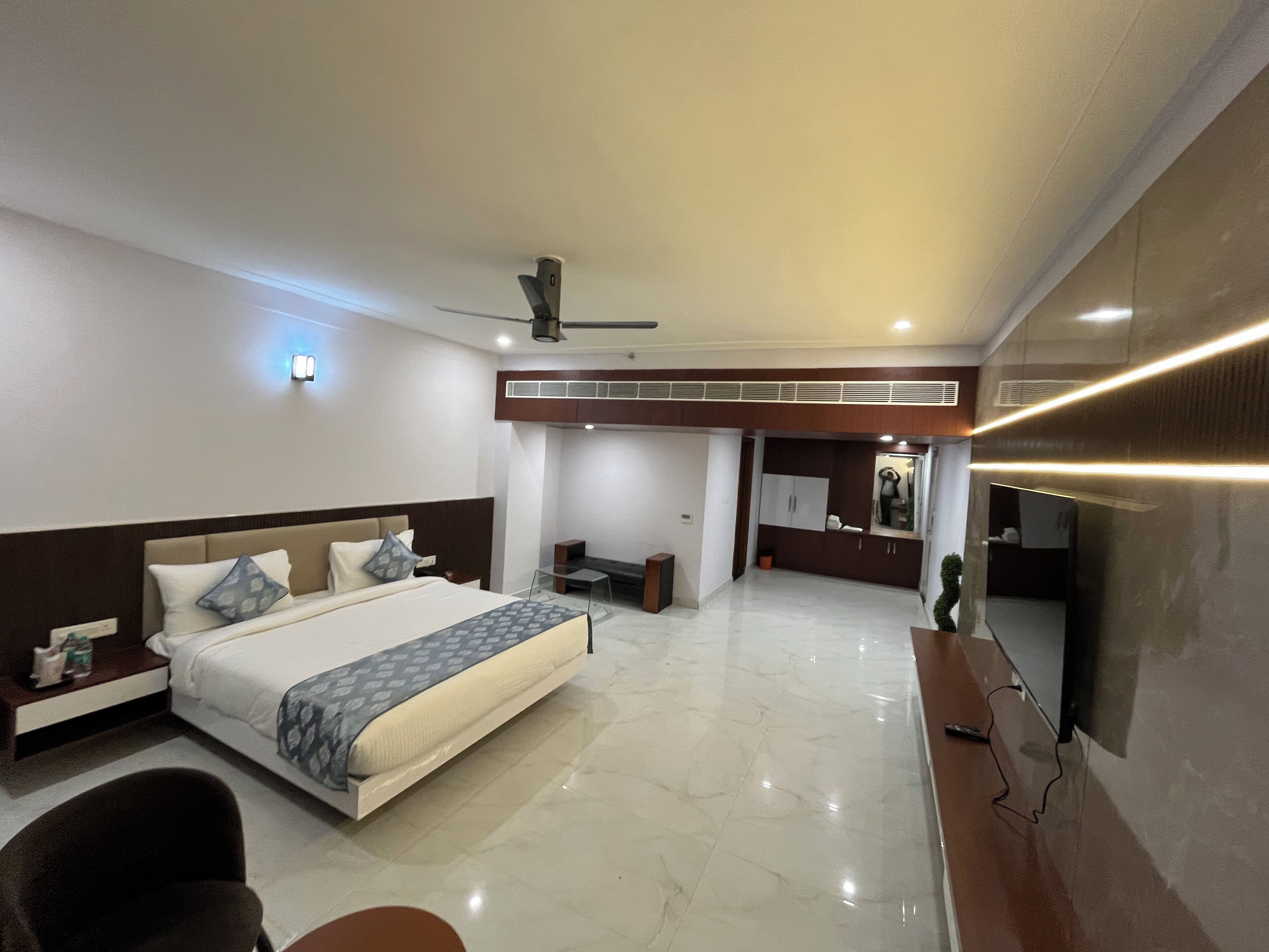 A modern hotel room with a large bed, a desk, a chair, a TV, and a ceiling fan.