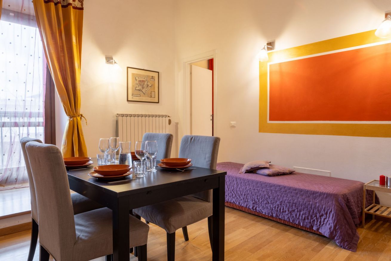 Een kamer met een eettafel gedekt voor vier personen, een bed met een paars dekbed en een groot oranje kunstwerk aan de muur.