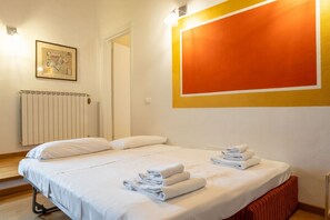 A hotel room with a bed, white linens, a radiator, a framed picture, and a large orange wall decoration. - Sant'Ambrogio Apartments - Raffaello (Firenze)