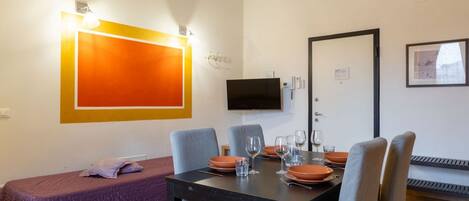 A modern room with a dining table set for four, a bed with a purple bedspread, a mounted TV, and a framed picture on the wall.