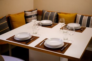 Una mesa de comedor dispuesta con platos blancos, copas de vino y cubiertos, rodeada por un sofá con cojines a rayas.