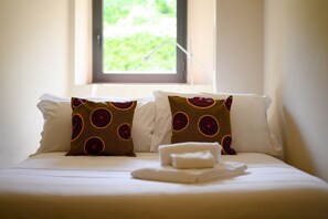 Una cama bien hecha con dos almohadas, una ventana con vistas a la vegetación y una mesita de noche con una toalla doblada.
