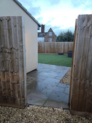 A patio area with a wooden fence, a paved walkway, and a grassy lawn. - The Nook | Cotswolds Home | HOT TUB | Sleeps 9 (Eastington)