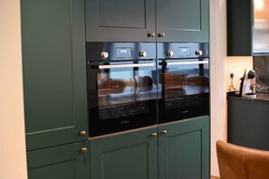 A kitchen with two Bosch ovens, green cabinetry, and a brown chair.