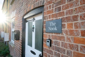 A brick building with a white door, a sign reading 'The Nook,' and a security device on the wall. - The Nook | Cotswolds Home | HOT TUB | Sleeps 9 (Eastington)