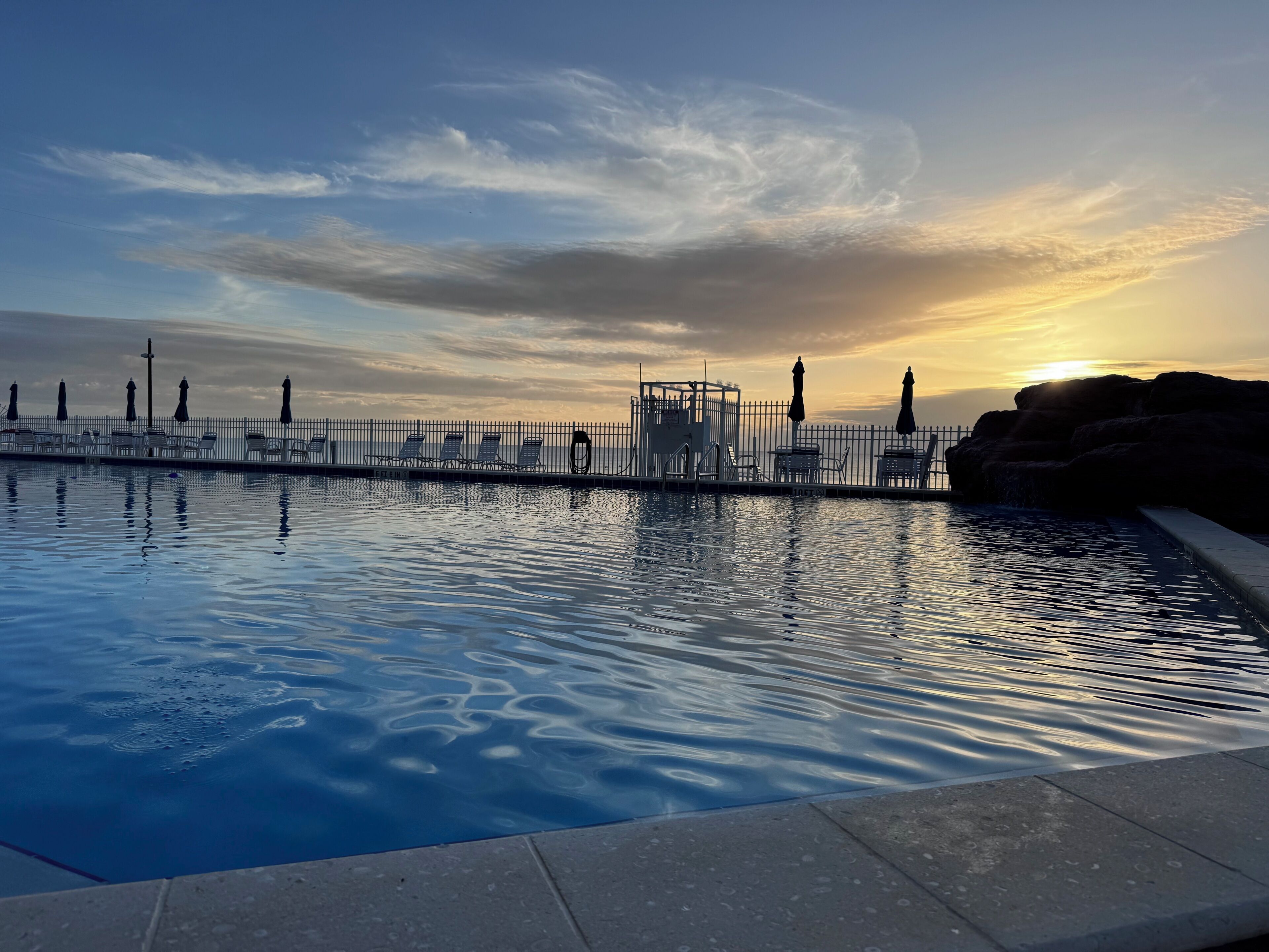 En swimmingpool med udsigt til solnedgang, et hegn og liggestole.