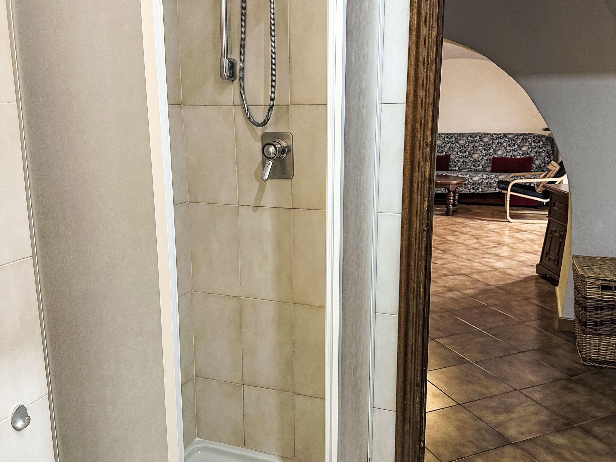 Un baño con azulejos color beige, una ducha con cabezal de mano y una puerta visible que da a una sala con un sofá estampado y una mesita redonda pequeña.