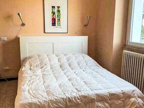 Une chambre à coucher avec une tête de lit blanche, un lit recouvert d’une couette blanche et un tableau encadré accroché au mur.