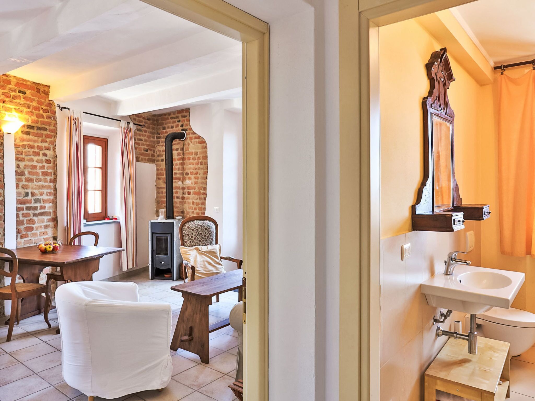 A room with a brick wall, a wooden table, a white chair, a sofa, a fireplace, and a bathroom with a mirror and sink.