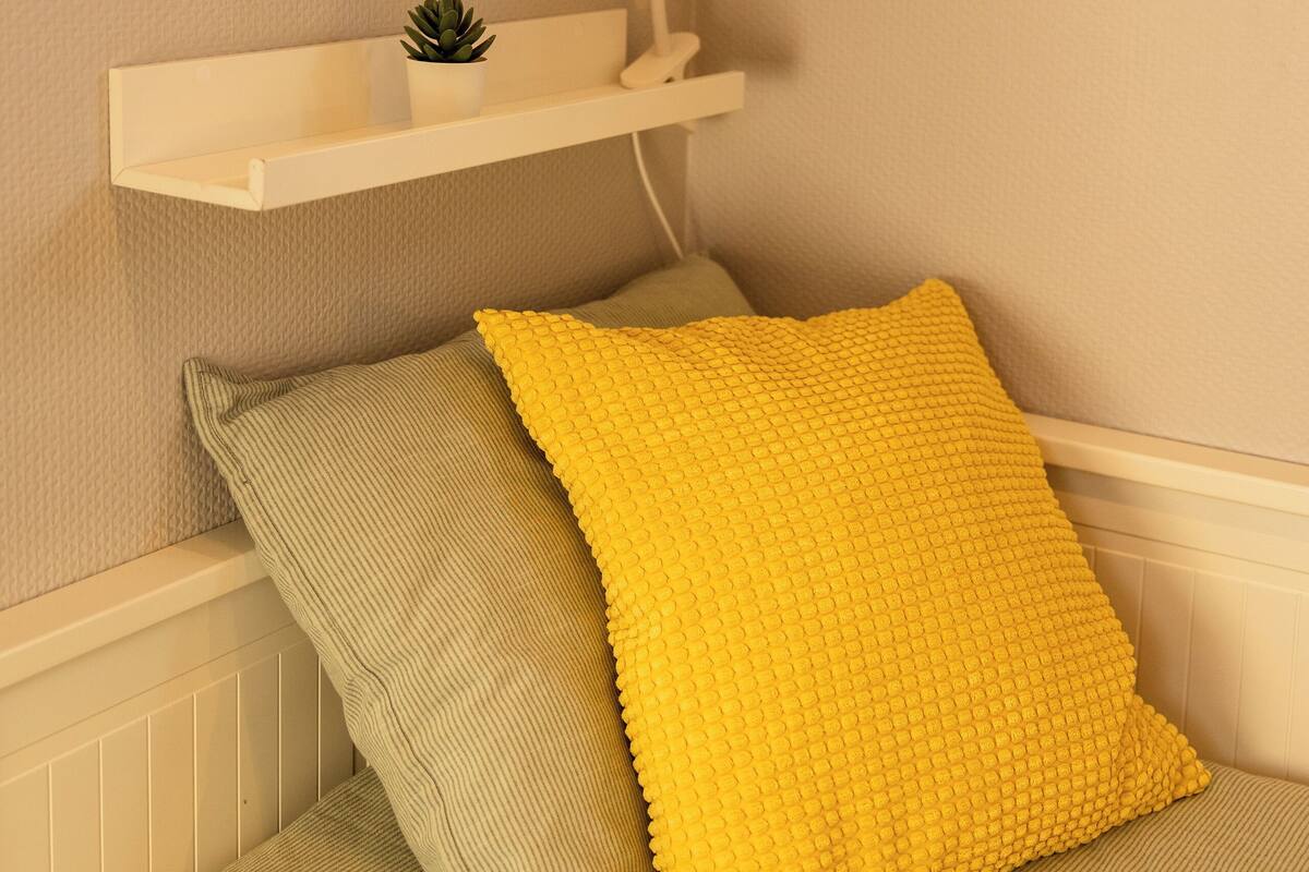 Eine Ecke des Raumes zeigt ein weißes Regal mit einem gerahmten Pflanzenprint, einem kleinen Topfpflanzen und einem gelben, strukturierten Kissen auf einer grünen Couch.