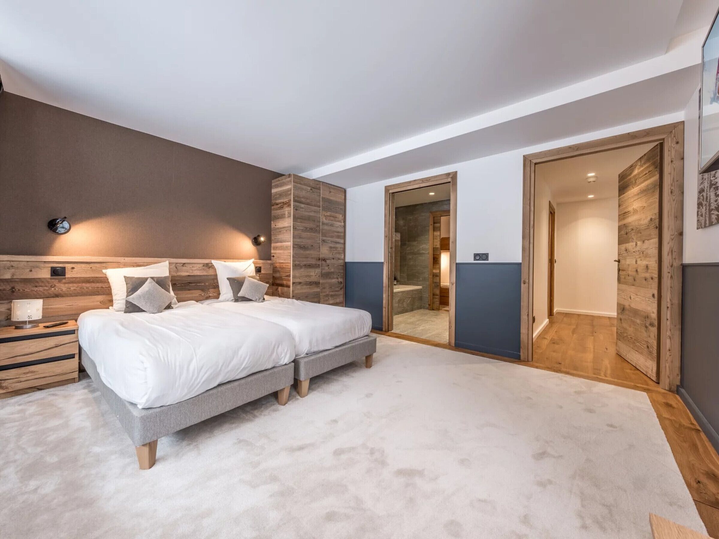 Een moderne slaapkamer met een groot bed, een nachtkastje en een houten hoofdbord. Er is een deur die leidt naar een badkamer en een gang.